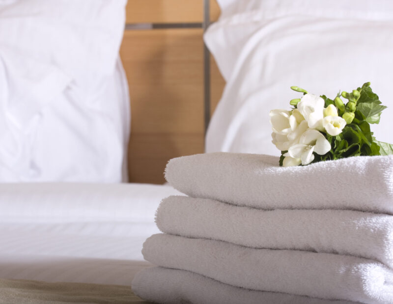 Hotel room with freshly made bed, perfectly clean and ironed snow white sheets, stack of new folded towels in natural sun light. Close up, copy space for text. High quality photo